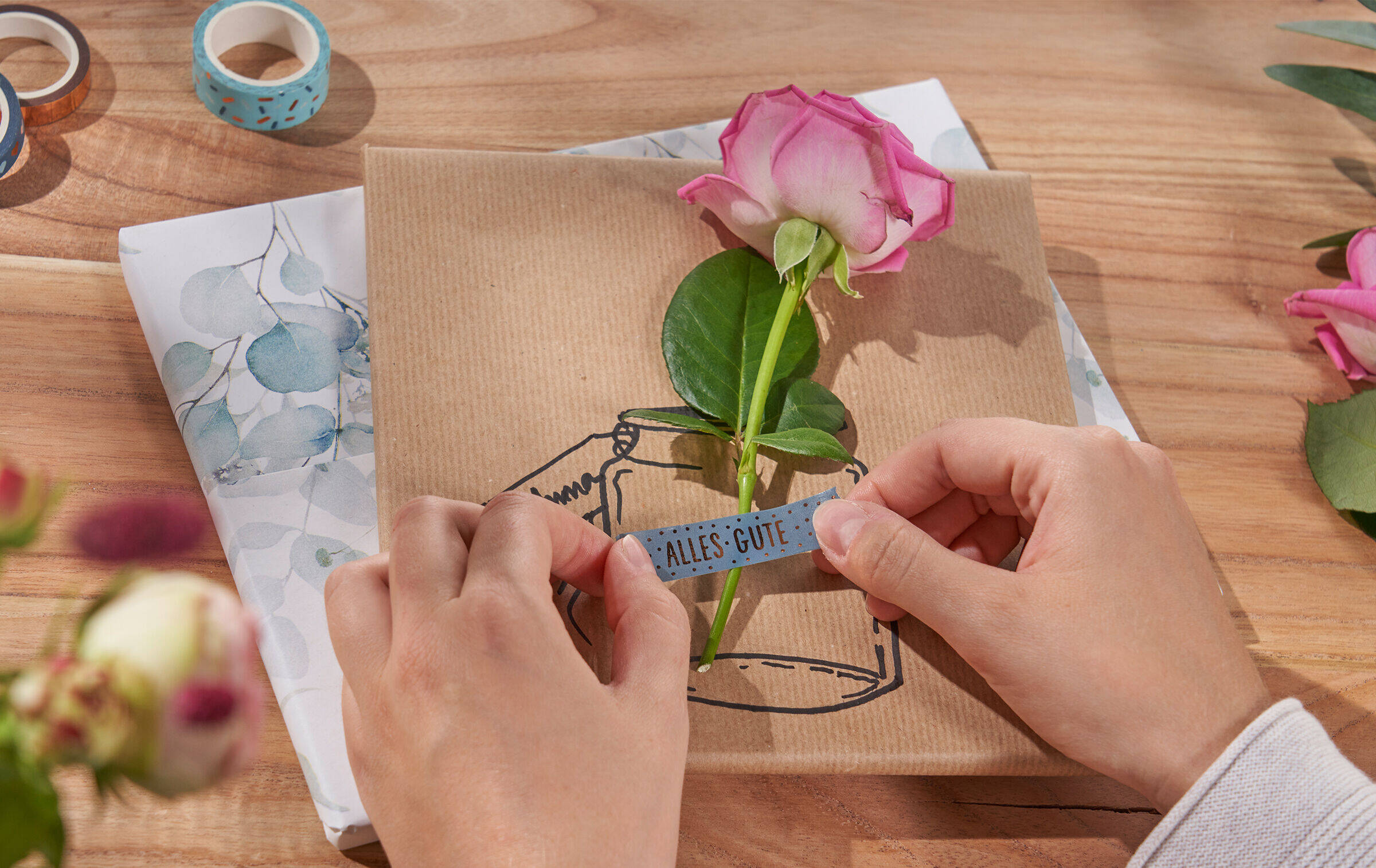 Zwei Hände kleben mit einem Stück Masking-Tape, auf dem „Alles Gute“ steht, eine Rose auf ein Geschenk, auf das eine Vasenillustration gezeichnet ist. Daneben lieben drei Rollen Masking-Tape und weitere Blumen.