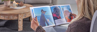 Eine blonde Frau hält ein CEWE FOTOBUCH in den Händen. Auf der Doppelseite sind Fotos von Kindern beim Bootfahren auf einem See zu sehen. Im Hintergrund ist ein graues Sofa und ein Holztisch sichtbar.