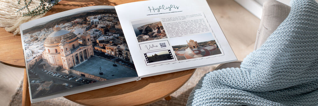 Ein aufgeschlagenes CEWE FOTOBUCH liegt auf einem Tisch neben einem Sofa. Auf der linken Seite ist ein Foto von Maltas Architektur abgebildet, auf der rechten Seite ist zusätzlich ein Video per QR-Code eingebunden.