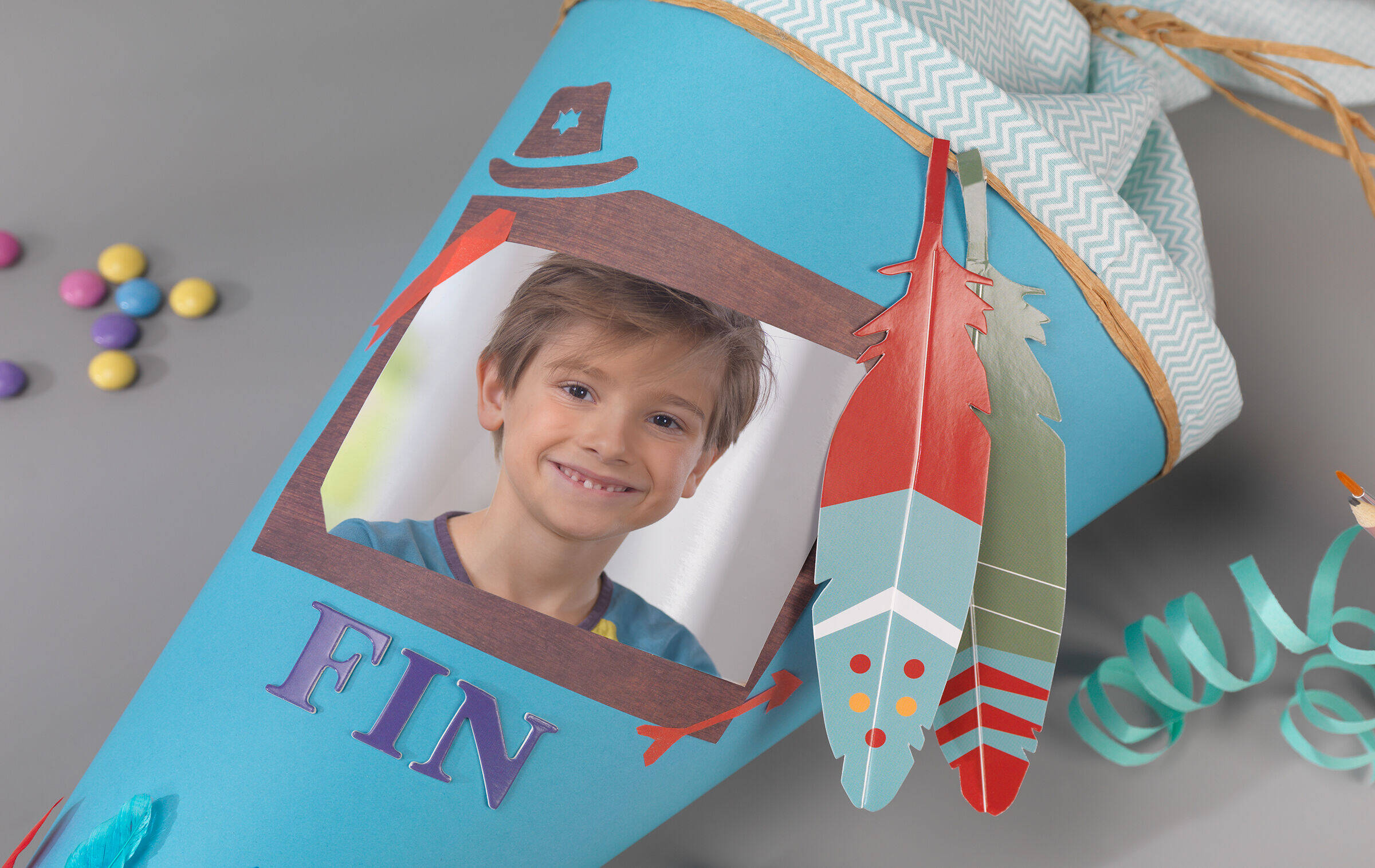 Die Detailaufnahme einer hellblauen Schultüte zeigt einen Fotosticker mit dem Motiv eines Jungen, das von bunten Papierfedern und dem Namen "Fin" eingerahmt wird.