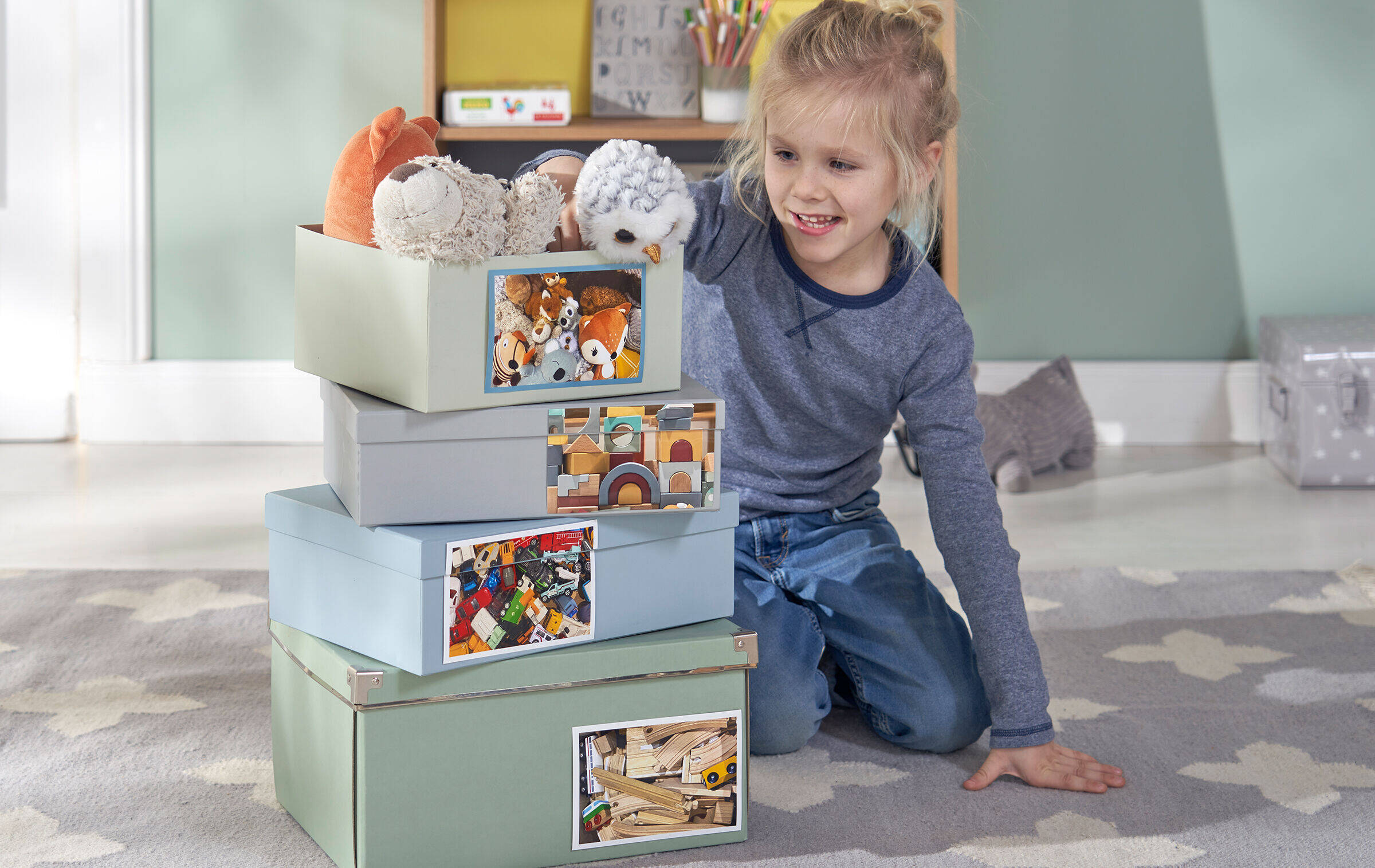 Kind kniet neben vier übereinandergestapelten Boxen. Darauf sind Fotos vom jeweiligen Inhalt der Boxen zu sehen, zum Beispiel Spielzeug oder Stofftiere.
