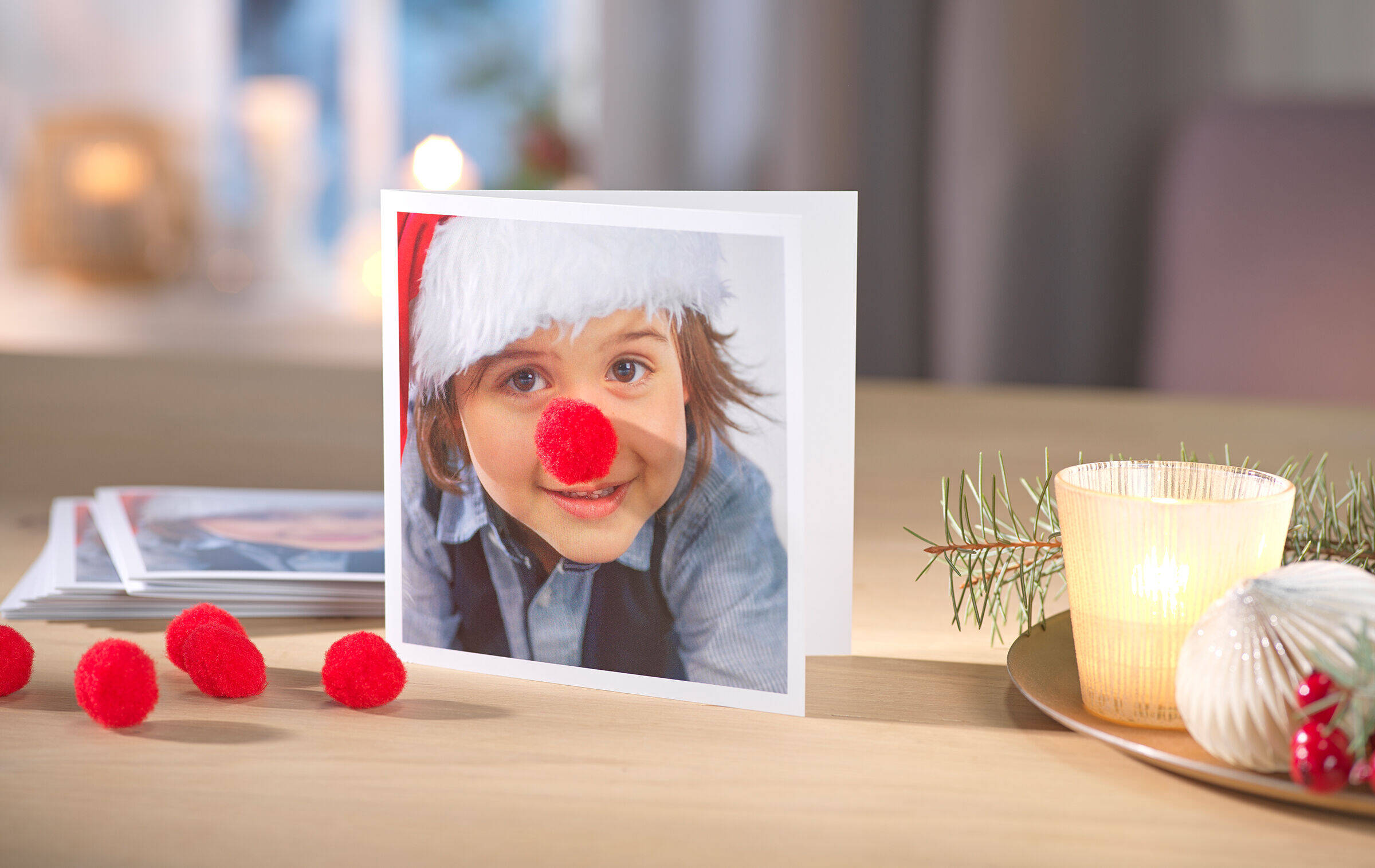 Weihnachtskarte mit Foto eines kleinen Kindes. Auf dessen Näschen wurde eine flauschige, rote Bommelnase geklebt.