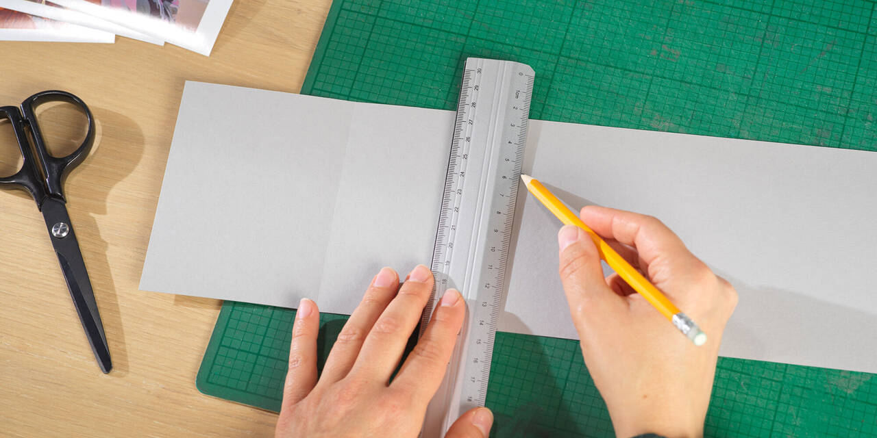 Zwei Hände zeichnen mit Bleistift und Lineal Abstände für die Fotos auf einen grauen Pappkarton.