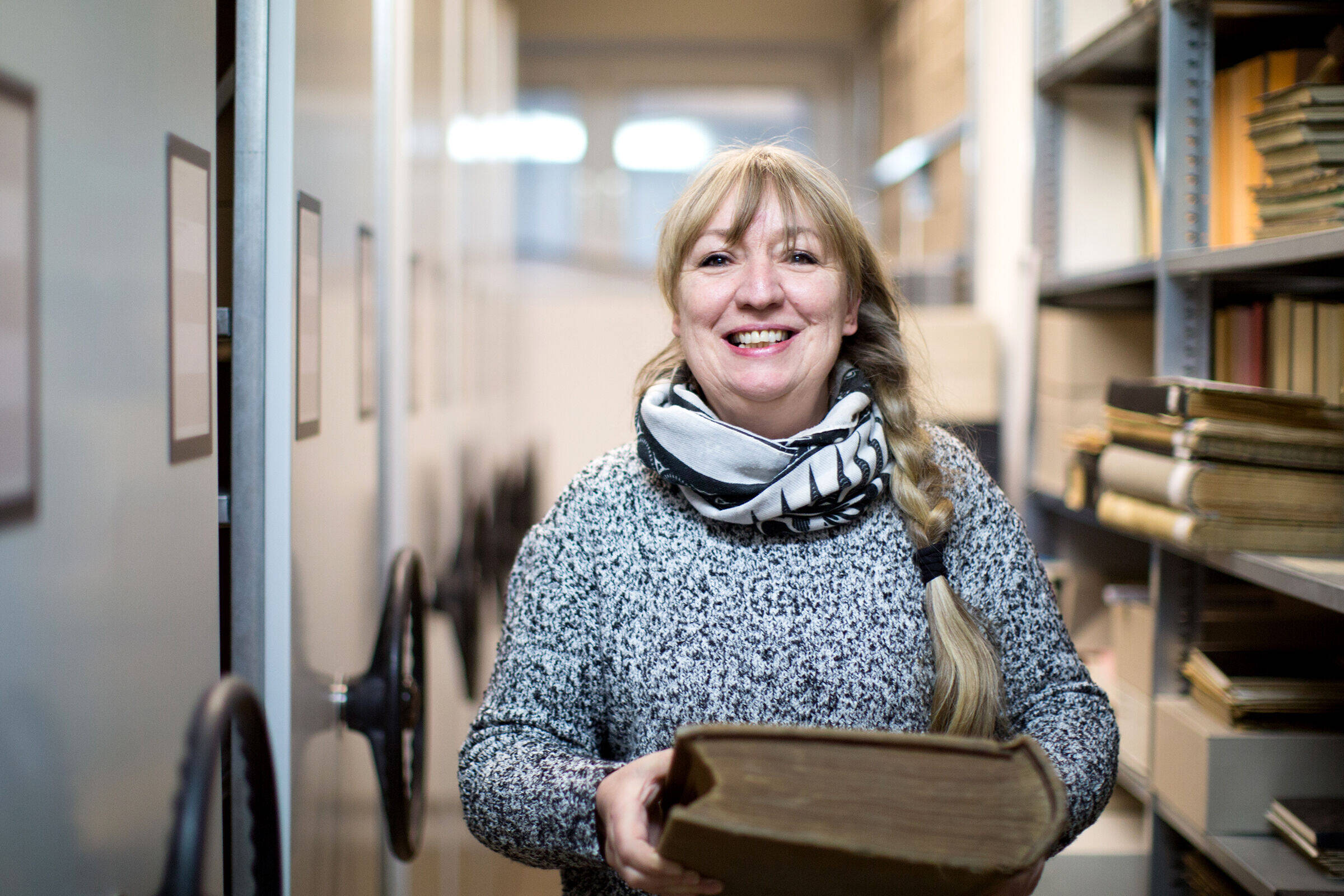 Archivarin Menna Hensmann steht in einem der Gänge des Leeraner Stadtarchivs. In den Händen hält sie ein älteres Buch. Dabei schaut sie lächelnd in die Kamera.