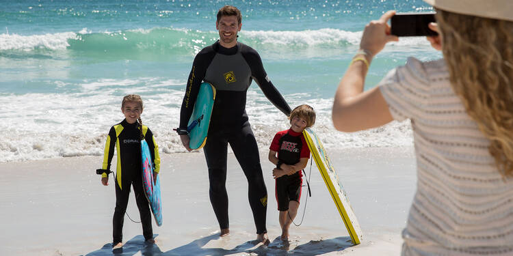 Zu sehen ist eine Familie am Strand. Eine Frau steht mit dem Rücken zur Kamera und fotografiert mit einem Smartphone ihren Mann und zwei Kinder, die mit Surfbrettern aus der Brandung kommen.