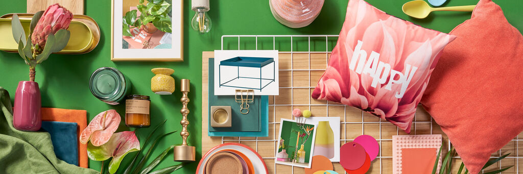 Das Foto zeigt ein Moodboard im Happy-Stil. Auf einem grünen Untergrund liegen verschiedene Accessoires wie Kerzenständer, Teller, Besteck und Bilderrahmen in unterschiedlichen bunten Farben.