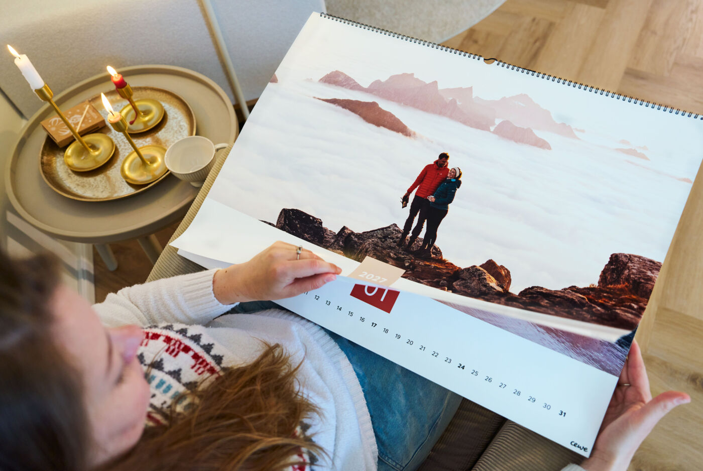 Frau sitzt in einem Sessel; neben ihr steht ein Tisch mit Kerzen, auf ihrem Schoß liegt ein Kalender.
