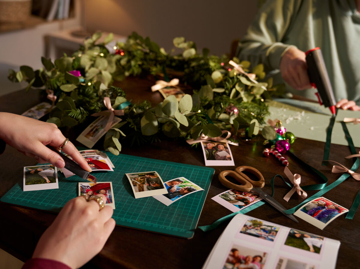Zwei Frauen basteln an einem Tisch, auf dem ein weihnachtlicher Kranz liegt sowie ausgedruckte Fotos und Bastelmaterial.