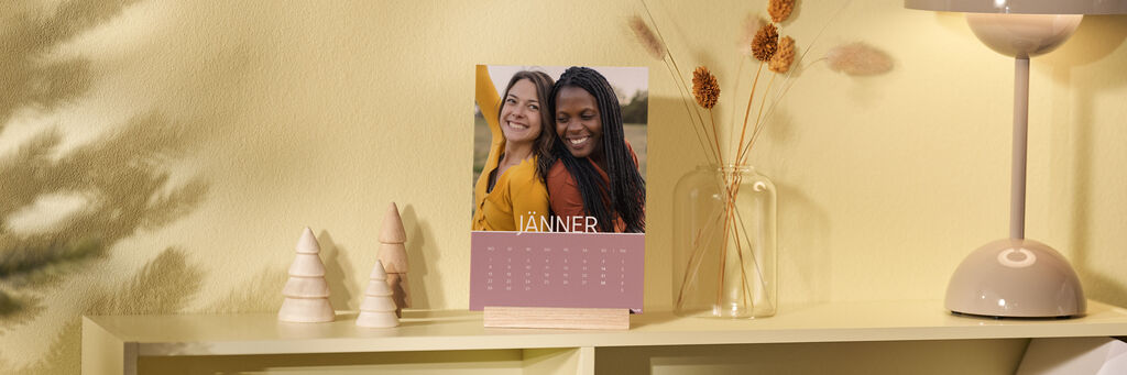 Ein Tischkalender Nature steht auf einem Regal. Das Kalenderblatt zeigt den Januar. Das Kalendarium hat einen rosa Hintergrund, darüber ist ein Foto von zwei Frauen zu sehen. Neben dem Kalender stehen weihnachtliche Holzdeko, eine Lampe und eine Vase.