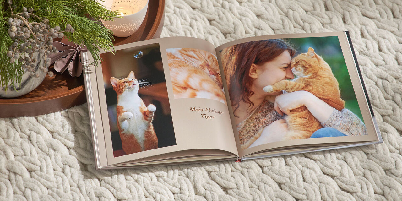 Ein aufgeschlagenes CEWE FOTOBUCH im quadratischen Format liegt auf einer weißen Decke. Auf der Doppelseite sind Fotos einer roten Katze und einer Frau zu sehen. Links neben dem Fotobuch liegt ein Tablett mit weihnachtlicher Deko und Kerzen.