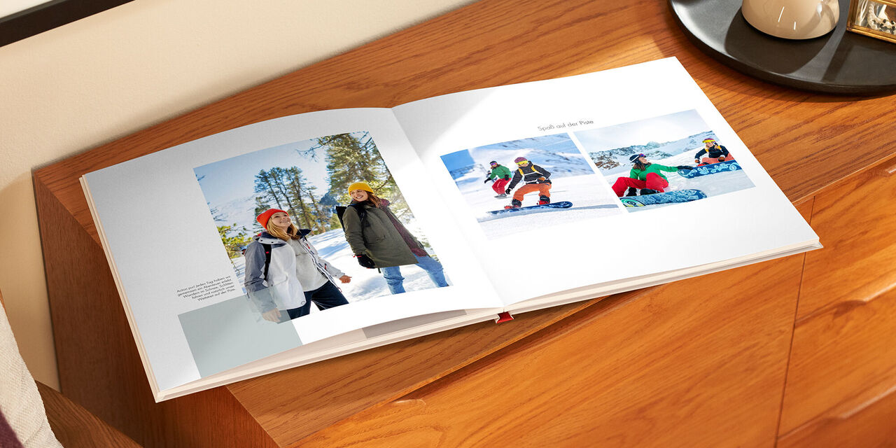 Auf einem Holzschrank liegt das aufgeklappte Fotobuch. Links sieht man ein Foto von zwei Frauen bei einer Wanderung. Daneben befindet sich ein kleiner Textblock. Auf der rechten Seite sind zwei Fotos der Frauen beim Snowboarden zu sehen. Darüber steht die Überschrift „Spaß auf der Piste“.