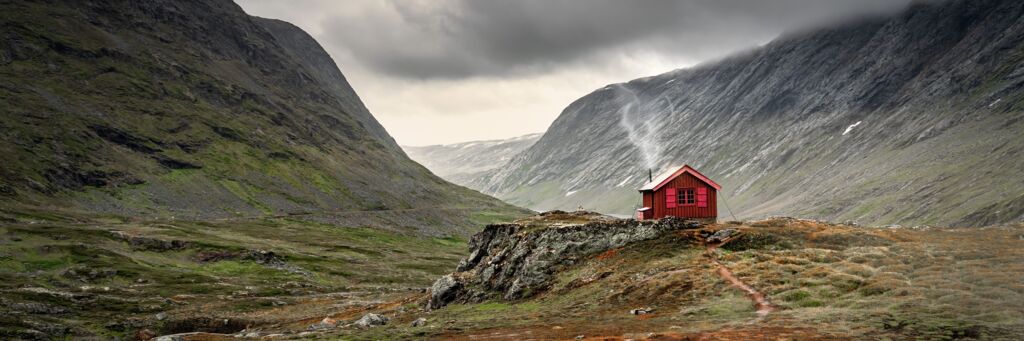 Rote Holzhütte auf einer Anhöhe in einem kargen, von steilen grauen Bergen umgebenen Tal unter einem bewölkten Himmel.