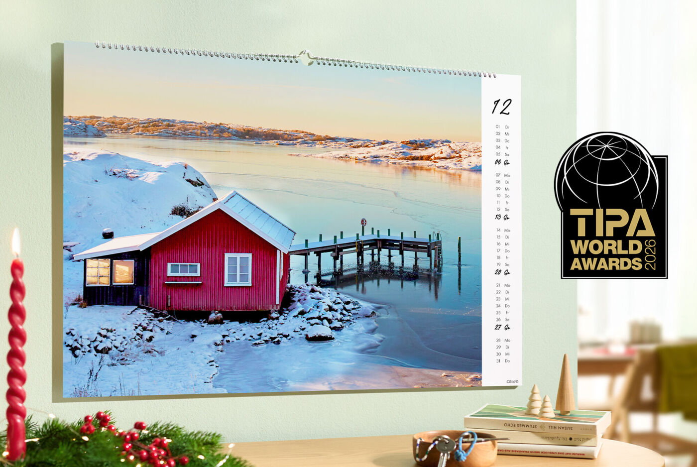 CEWE Kalender mit dem Bild eines roten Hauses an einem verschneiten See hängt an einer Wand in weihnachtlicher Umgebung, TIPA World Awards