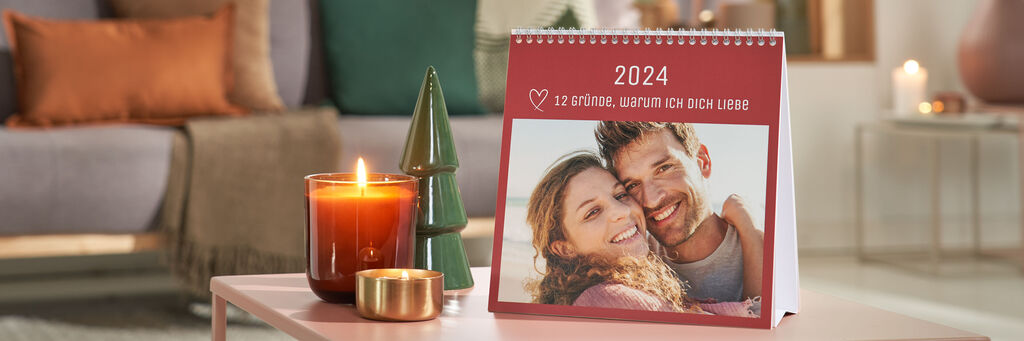 Ein Tischkalender mit dem Foto eines Liebespaares steht auf einem weihnachtlich dekorierten Wohnzimmertisch. Darauf sind das Foto eines Paares und darüber der Text „2022 – 12 Gründe, warum ich Dich liebe“ zu sehen.