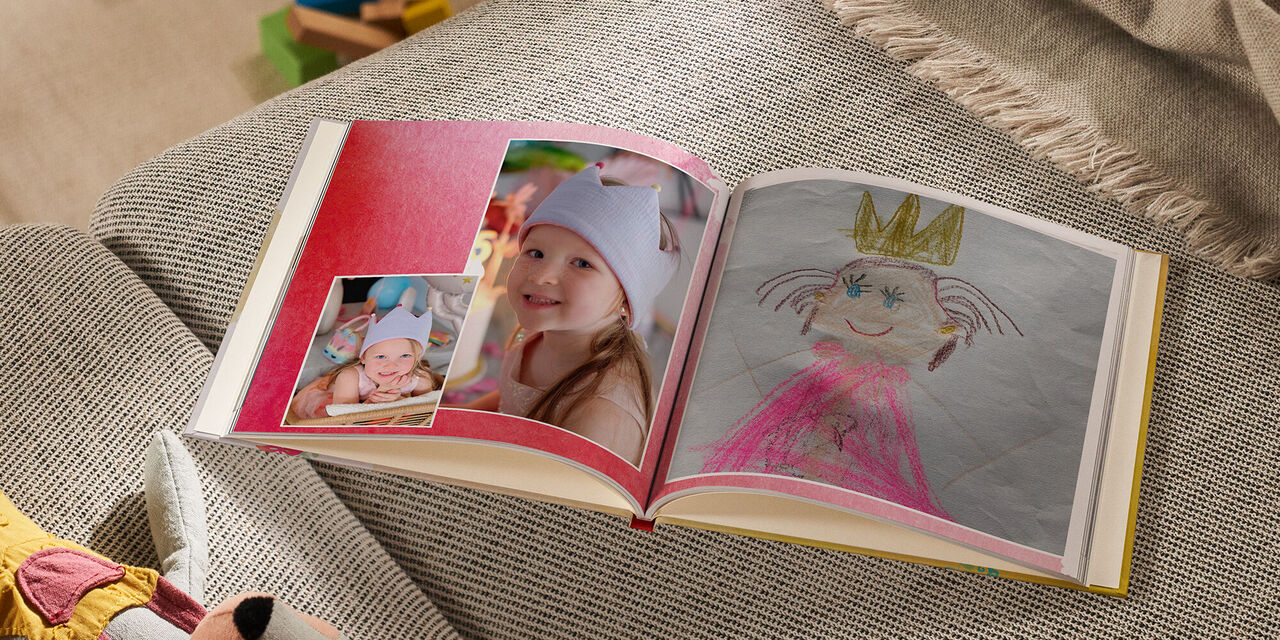 Auf einem Sofa liegt ein geöffnetes CEWE FOTOBUCH neben einem Kuscheltier und einer Decke. Die linke Seite zeigt zwei Fotos von einem lächelnden Mädchen, das eine Krone trägt. Auf der rechten Seite ist eine Kinderzeichnung von einer Prinzessin mit einer Krone zu erkennen. Der Hintergrund der Doppelseite ist mit einem roten Aquarellmuster gestaltet.