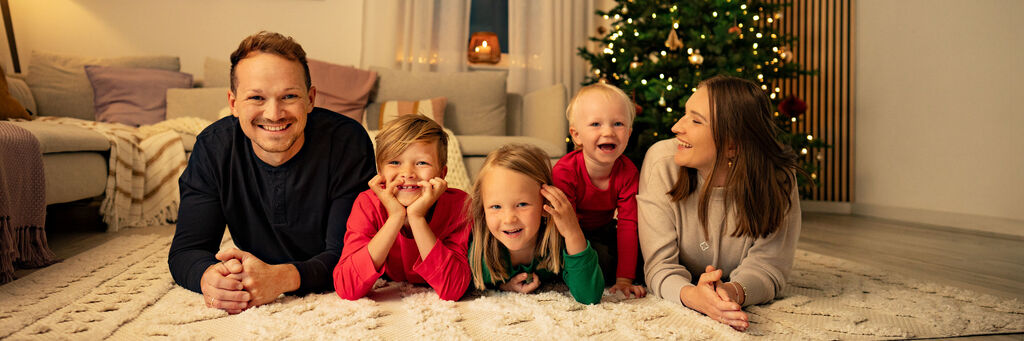 Eine fünfköpfige Familie liegt bäuchlings und auf die Ellenbogen aufgestützt, auf einem hellen Teppich und blickt in die Kamera. Die Kinder tragen gemütliche Kleidung in  
weihnachtlichen Farben. Vater und Mutter rahmen den Nachwuchs ein. Im Hintergrund ist ein weihnachtlich geschmücktes und beleuchtetes Wohnzimmer mit Tannenbaum zu sehen.