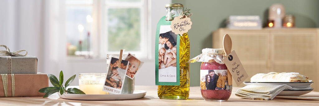 Auf einem Tisch stehen eine Kerze, ein Kräuteröl und ein Glas Marmelade. Alle Gefäße haben einen personalisierten Geschenkanhänger, auf denen Fotos einer Familie zu sehen sind.