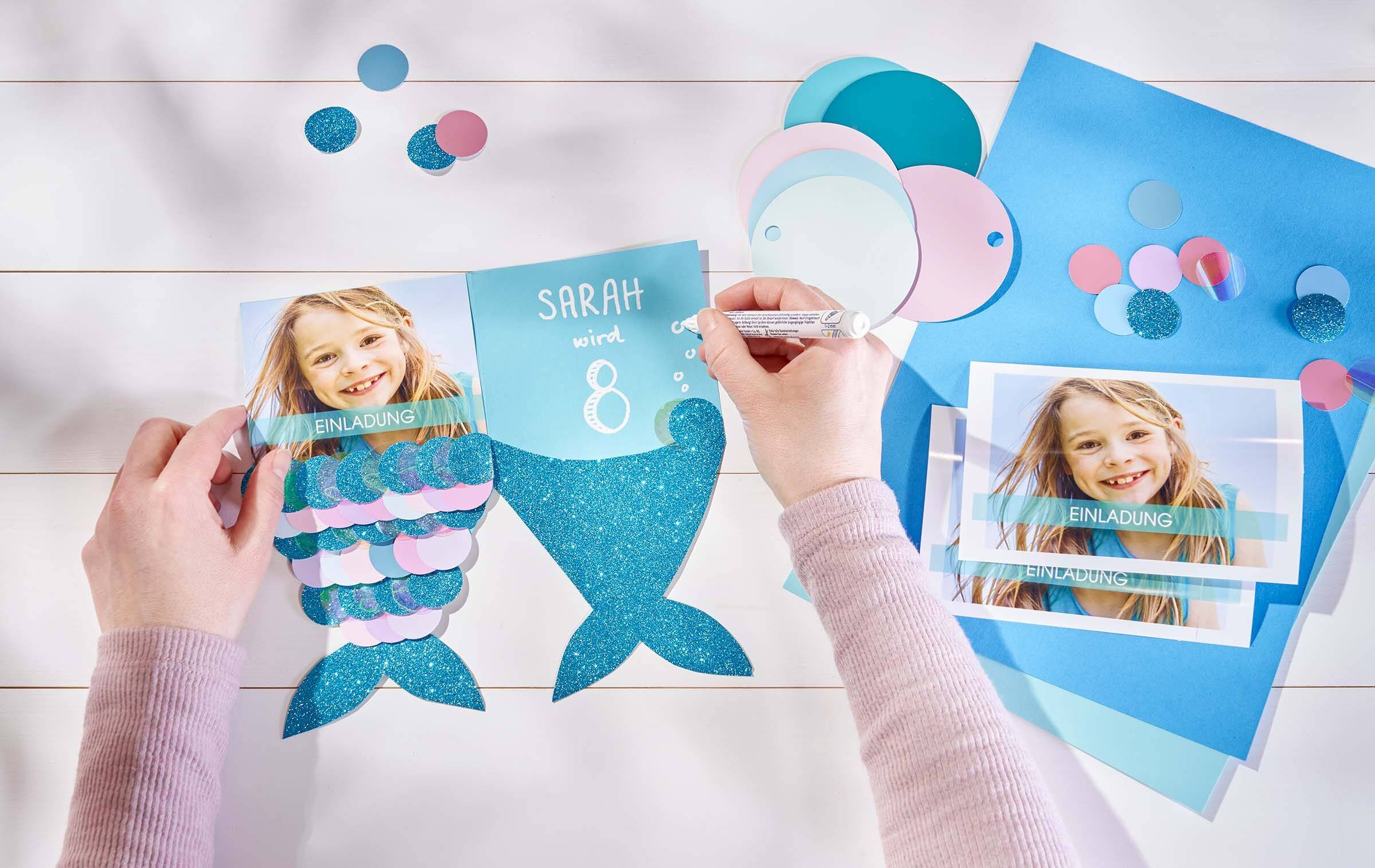 Zwei Hände beschriften eine Geburtstagseinladung im Meerjungfrauen-Stil. Auf der linken Seite ist ein Sofortfoto des Geburtstagskindes zu sehen. Im rechten Teil steht die Aufschrift “Sarah wird 8”. Daneben liegen weitere Fotos, Tonpapier und kleine Kreise zum Aufkleben.