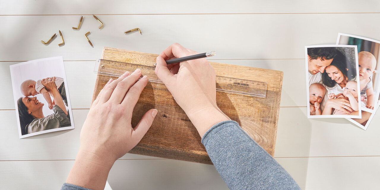 Nahaufnahme von zwei Händen, die mit Bleistift und Lineal Markierungen auf einem Holzblock einzeichnen. Daneben liegen CEWE Sofortfotos und Hakenschrauben.