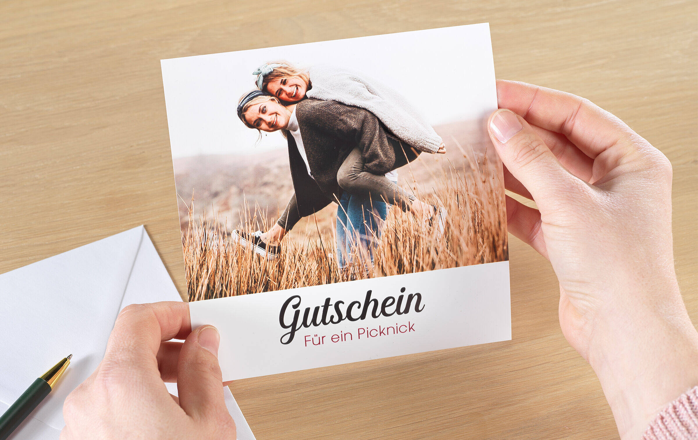 Ein Gutschein für ein Picknick ist mit dem Foto zweier Freundinnen bebildert und wird in Händen gehalten. Daneben liegen ein Briefumschlag und ein Stift.