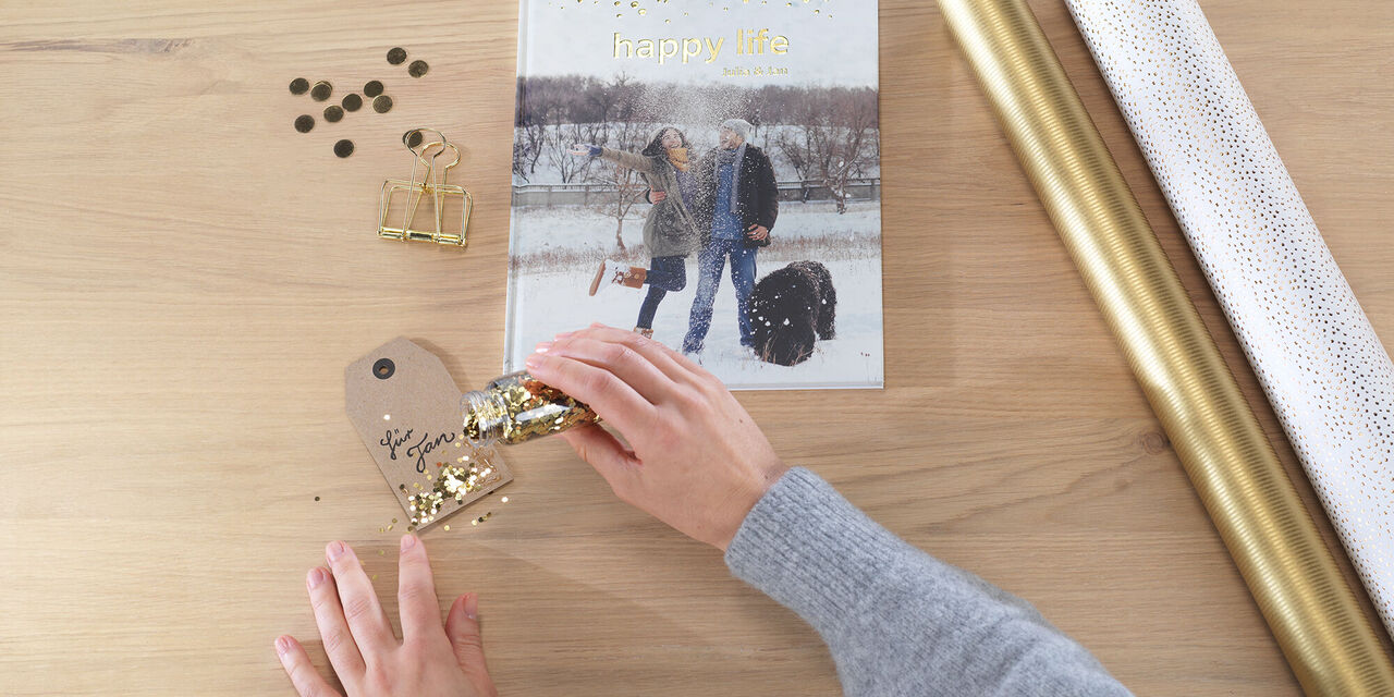 CEWE FOTOBUCH mit Goldveredelung liegt auf einer Tischplatte neben passendem Geschenkpapier. Zwei Hände streuen Goldglitzer auf die Geschenkkarte, auf der "Für Jan" steht.