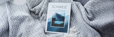 Ein CEWE FOTOBUCH zum Reiseziel Schweiz liegt auf einer Decke und einem Kissen neben einer Zimmerpflanze.