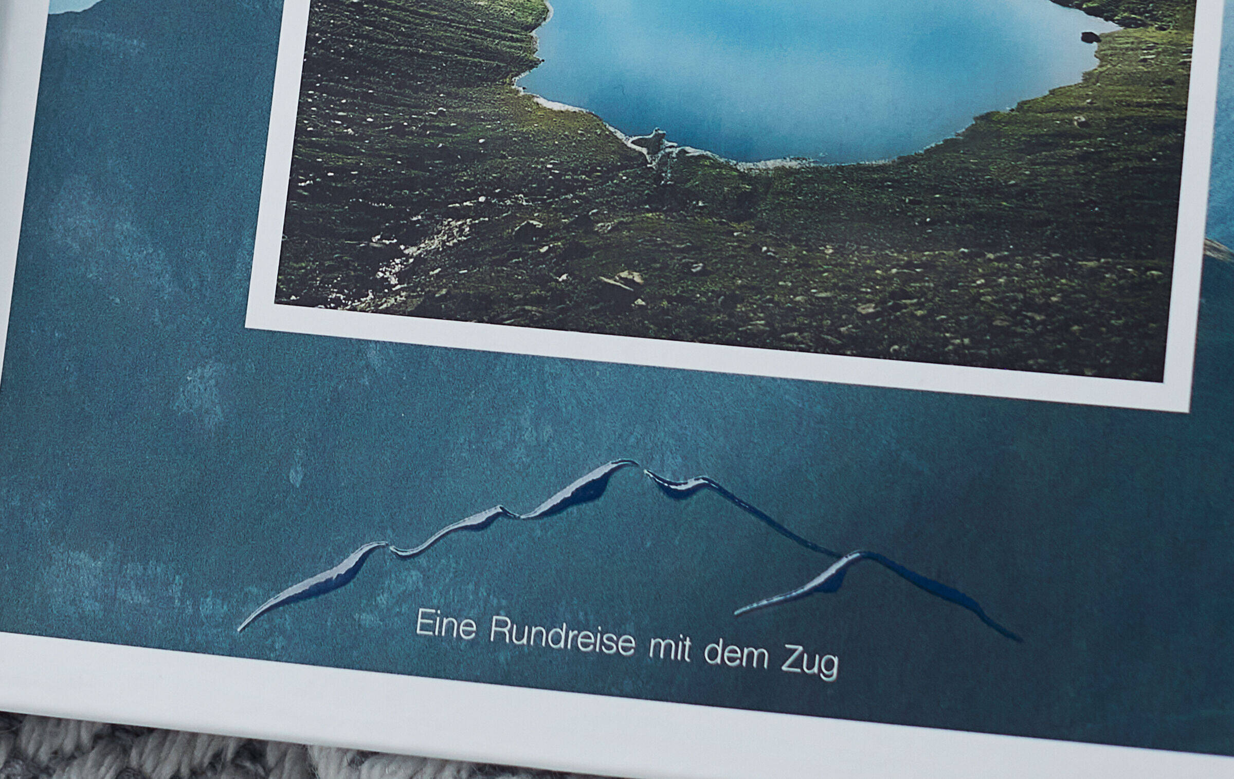 Zu sehen ist die Detailansicht einer schimmernden Clipart-Bergkontur auf dem Einband des Fotobuchs. Die Textzeile darunter lautet: "Eine Rundreise mit dem Zug".