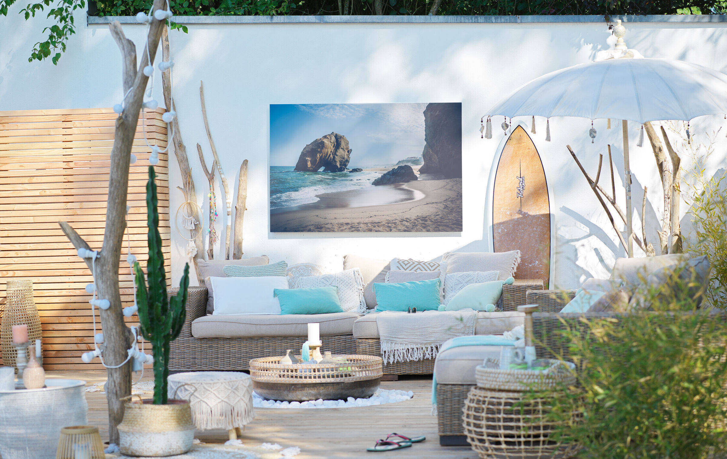 Großformatiges Wandbild mit Strandmotiv hängt an einer Gartenmauer am Rande einer Terrasse mit gemütlichen Lounge-Möbeln.