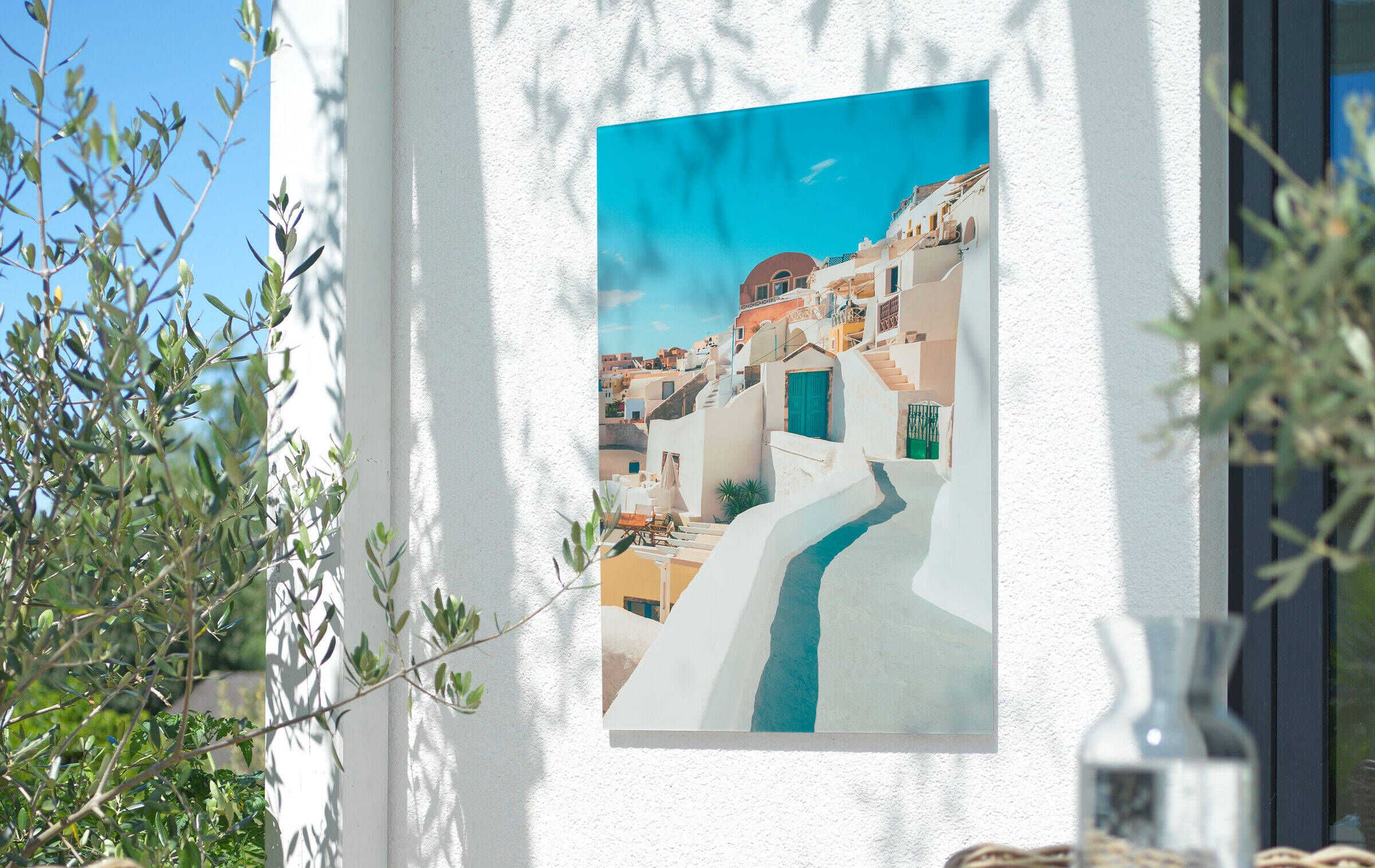 Wandbild mit mediterranem Stadtmotiv hängt an Hauswand auf einem Balkon. Im Vordergrund ist unscharf ein gedeckter Esstisch zu sehen.