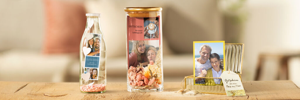 Auf einem Holztisch stehen drei Geschenke nebeneinander. Links eine Flasche mit Herzen und Sofortfotostreifen, in der Mitte ein Glas mit Trockenblumen und Collage und rechts eine Schatulle mit Sofortfoto und Sand.