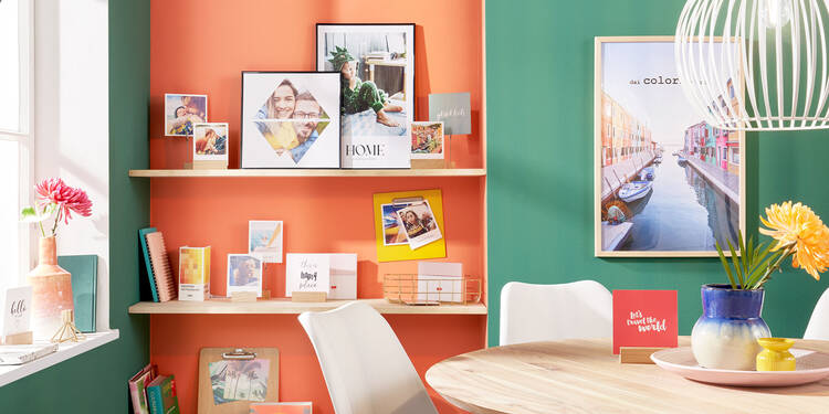 In einem Wohnraum steht ein Esstisch. Links dahinter ist eine Vertiefung in einer Wand sichtbar, die als Regal genutzt wird. Der Hintergrund der Nische ist Orange gestrichen, die restliche Wand grün. Auf den Regalbrettern stehen viele verschiedene Fotos, gerahmte Poster und Bücher.