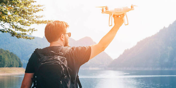Ein Mann steht an einem Bergsee, die Sonne scheint. In der rechten erhobenen Hand hält er eine Drohne fest.