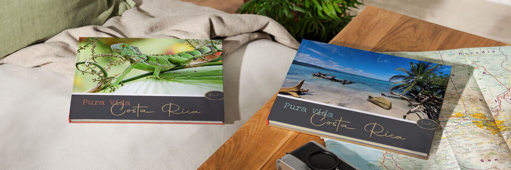 Zwei Fotobücher liegen zugeklappt auf einem Tisch und Sofa. Beide zeigen das Cover "Pura Vida Costa Rica". Auf der linken Seite liegt Teil 1 und zeigt einen Leguan. Auf der rechten Seite liegt Teil 2 und zeigt einen Strand.
