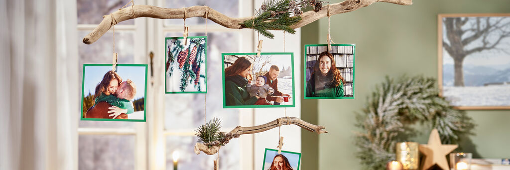 In einem Wohnzimmer hängt ein Fotomobile vor einem Fenster. Dafür wurden mithilfe von Bändern und Wäscheklammern aus Holz an zwei Ästen Sofortfotos mit grünen Rändern befestigt. Die Bilder zeigen Motive einer Familie mit Kind im Winter.