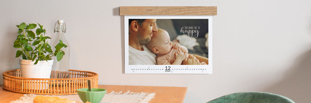 An einer Wand hängt über einem Tisch ein Wandkalender im Querformat mit Babymotiven. Er zeigt den Monat Dezember und hat eine Holzleiste, die die Ringbindung verdeckt.