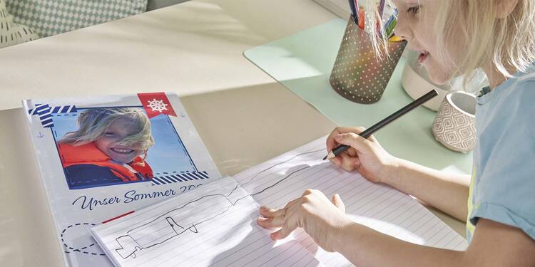 Ein kleiner Junge malt in einem linierten Notizbuch, das auf einem Tisch liegt. Daneben liegt ein weiteres Notizbuch auf dessen Einband ein Foto von einem zweiten Jungen zu sehen ist. Darunter steht „Unser Sommer“.