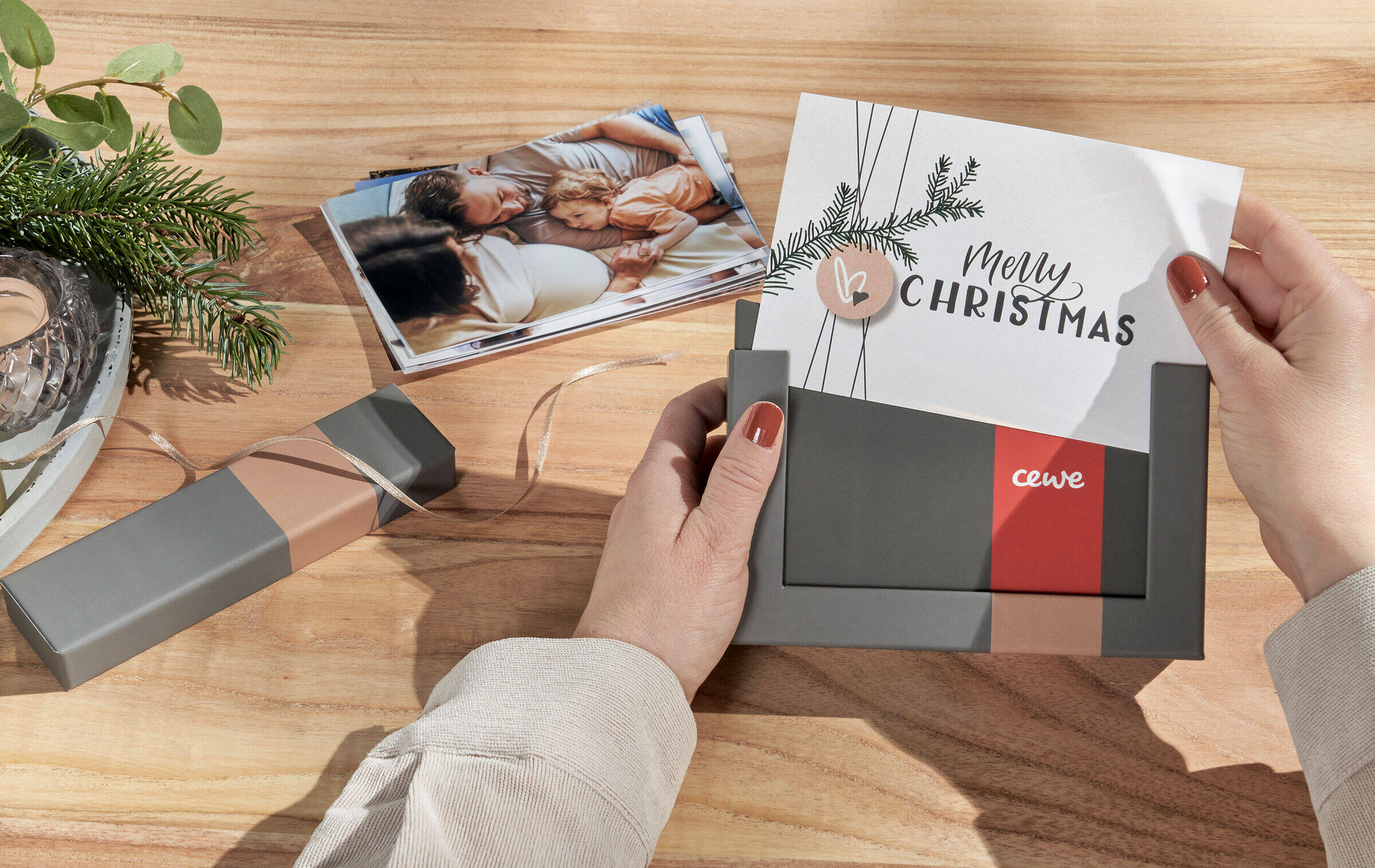 Zwei Hände schieben ein Bild, auf dem „Merry Christmas“ steht, in den Bilderrahmen der Bilderbox. Daneben steht ein weihnachtliches Gesteck und es liegt ein Stapel Familienfotos auf dem Tisch.